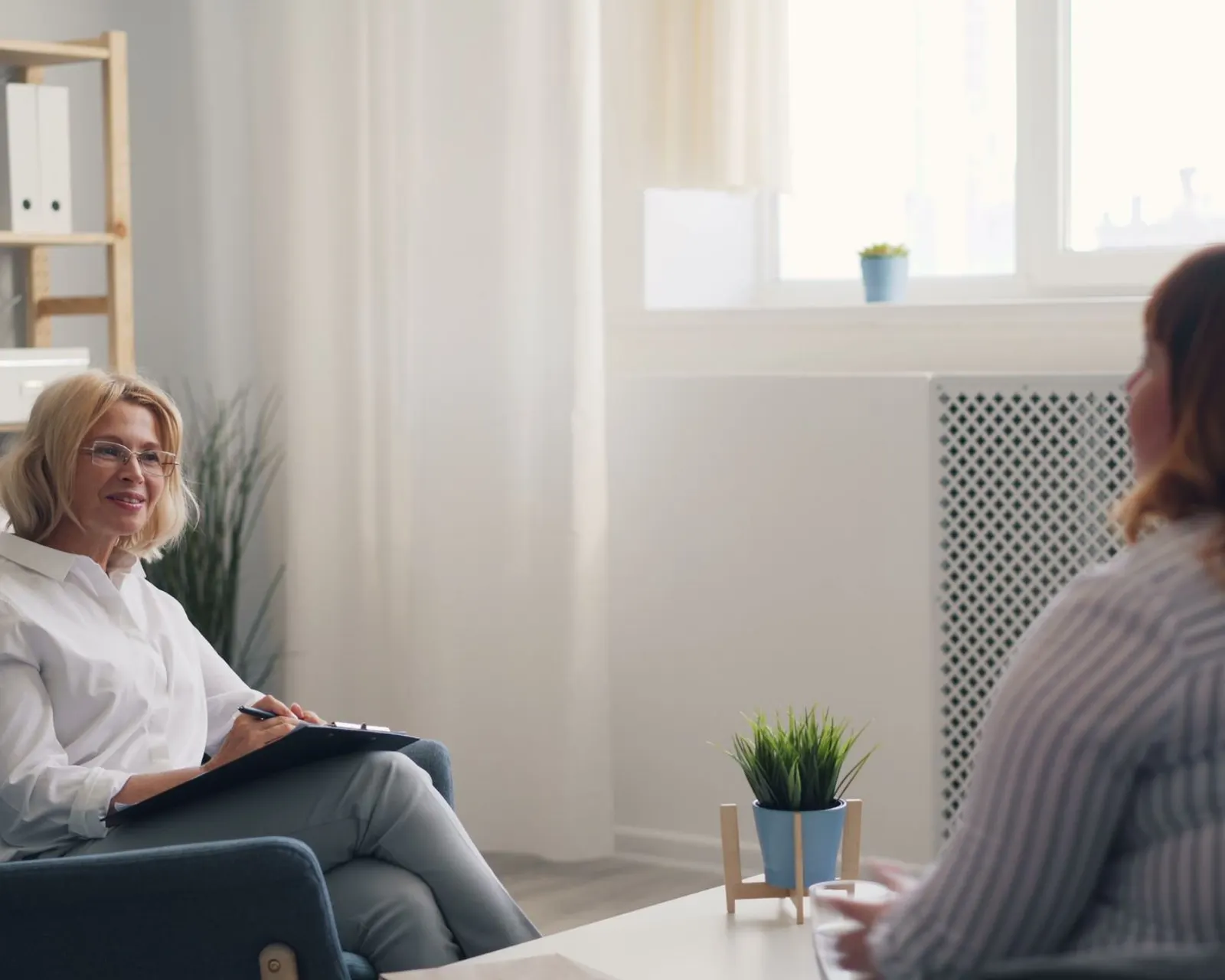 A Heilpraktiker sits opposite her patient in the consulting room, taking notes on a clipboard.